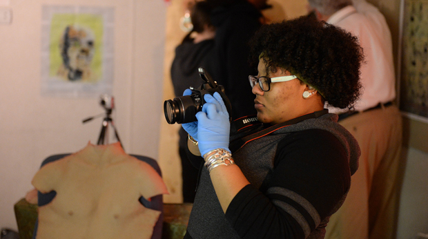 A student wearing blue gloves takes pictures with a camera in the crime scene investigation lab at La Roche University. 